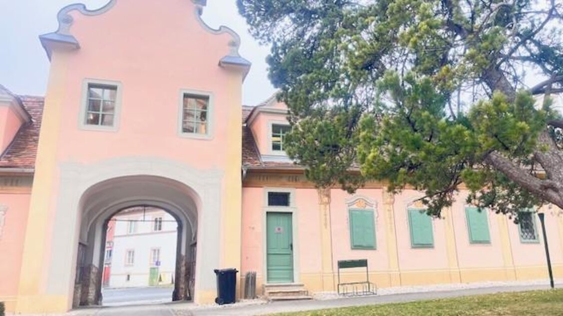 Ein historisches Gebäude mit einer rosa Fassade und einem Torbogen. Rechts ist ein großer Baum und eine Bank im Vordergrund. | © Tourismusverband Oststeiermark