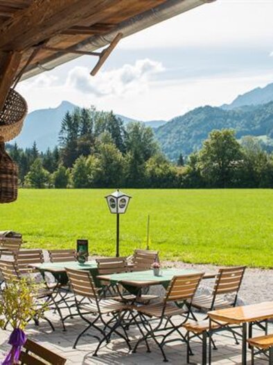Ein einladendes Restaurant mit Holzterrasse und Tischen im Freien. Im Hintergrund ist eine malerische Landschaft mit grünen Wiesen und Bergen zu sehen.