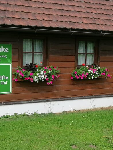 Ein Holzhaus mit drei Fenstern und dekorativen Blumenkästen. Es gibt ein Schild, das Getränke und Wein zum Selbstbedienen anbietet. | © Fam. Kirchleitner