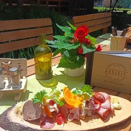 Tisch gedeckt im Außenbereich mit einem Holzbrett, auf dem eine Auswahl an Aufschnitt, Käse und frischem Gemüse serviert wird. Im Hintergrund ein Menübuch mit der Aufschrift „AUSZEIT“ und eine Flasche Öl auf einem Tisch unter Sonnenschein. | © Kager's Auszeit