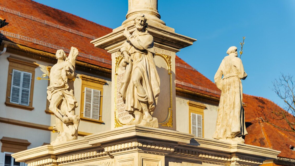 Steinerne Säule mit drei Heiligenfiguren vor einem historischen Gebäude mit rotem Ziegeldach unter blauem Himmel in Bad Radkersburg. | © Zehnerhaus Bad Radkersburg