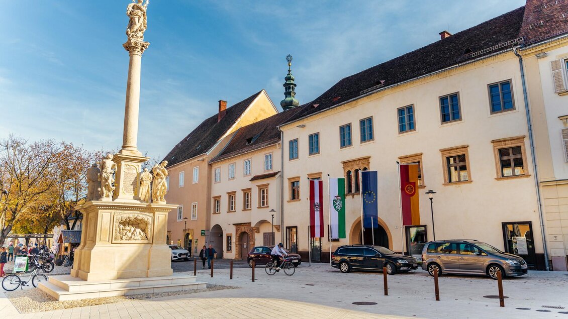 Hohe Mariensäule mit Figuren auf einem historischen Platz, umgeben von alten Gebäuden und herbstlichen Bäumen. | © Zehnerhaus Bad Radkersburg