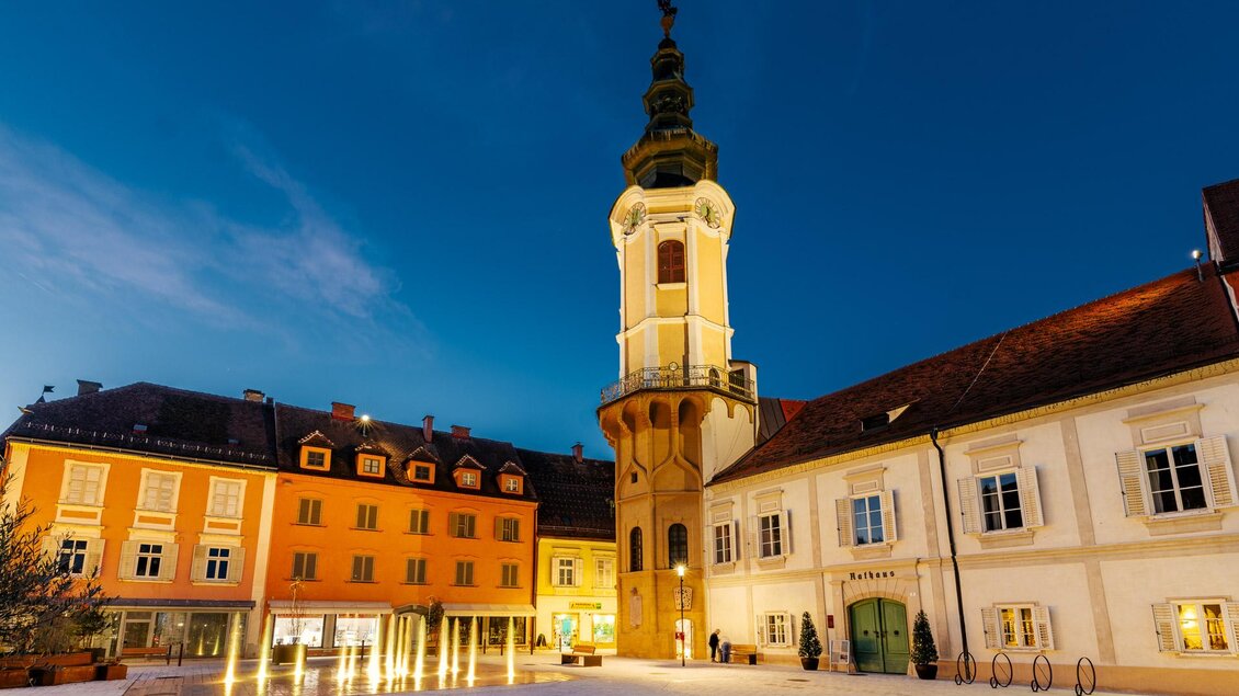 Rathausplatz in Bad Radkersburg bei Abenddämmerung mit historischem Rathausturm, beleuchteten Bürgerhäusern und Wasserspiel in der Altstadt. | © Zehnerhaus Bad Radkersburg