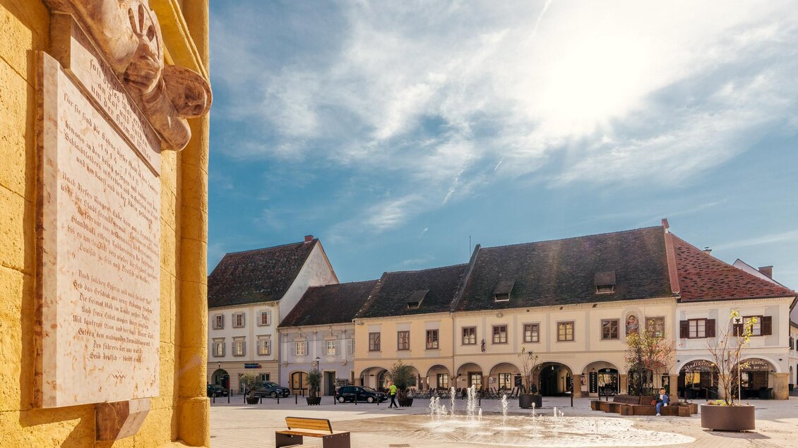 Sonniger Stadtplatz mit Wasserspiel, Sitzbänken und historischen Häusern mit Arkaden. Links ist eine steinerne Gedenktafel zu sehen. | © Zehnerhaus Bad Radkersburg