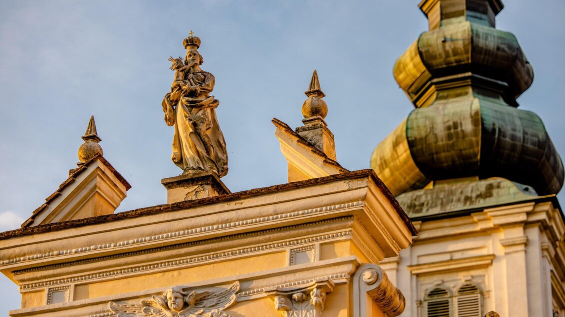 Detailansicht einer historischen Kirchenfassade mit Marienfigur und Kind, reich verzierten Bauelementen und Kirchturm im Hintergrund. | © Zehnerhaus Bad Radkersburg