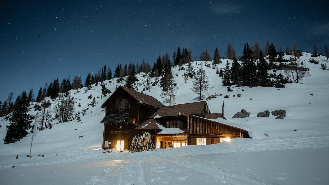 Gemütliche Berghütte im Winter bei Nacht, umgeben von Schnee und beleuchtetem Innenraum. | © René Sendlhofer-Schag