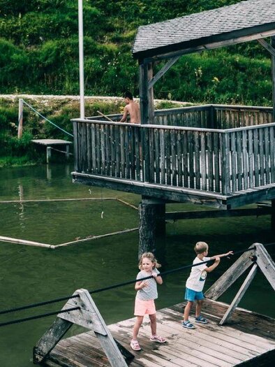 Two children are playing on a wooden walkway near a body of water. In the background, a wooden house on stilts stands. | © Armin Walcher