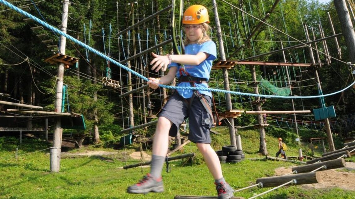Ein Kind mit einem Helm balanciert auf einem Seil im Abenteuerpark. Im Hintergrund sind Kletterstrukturen und Bäume zu sehen. | © Freelife Outdoorsport GmbH