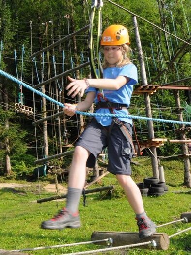 Ein Kind mit einem Helm balanciert auf einem Seil im Abenteuerpark. Im Hintergrund sind Kletterstrukturen und Bäume zu sehen. | © Freelife Outdoorsport GmbH