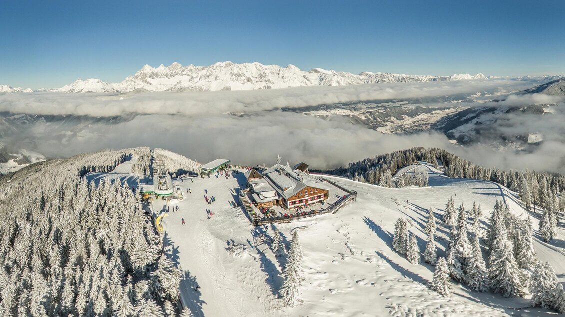 Eine schneebedeckte Berglandschaft mit einer Berghütte und vielen Tannenbäumen. Im Hintergrund sind hohe Berge und ein klarer Himmel zu sehen. | © Bence Bankuty