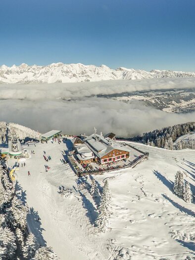 Eine schneebedeckte Berglandschaft mit einer Berghütte und vielen Tannenbäumen. Im Hintergrund sind hohe Berge und ein klarer Himmel zu sehen. | © Bence Bankuty