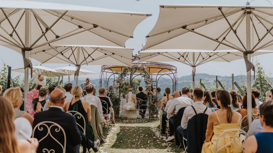 Eine elegante Hochzeitszeremonie im Freien mit Sonnenschirmen. Gäste sitzen in einem Garten, um einen festlichen Moment zu erleben. | © Weingartenhotel Harkamp