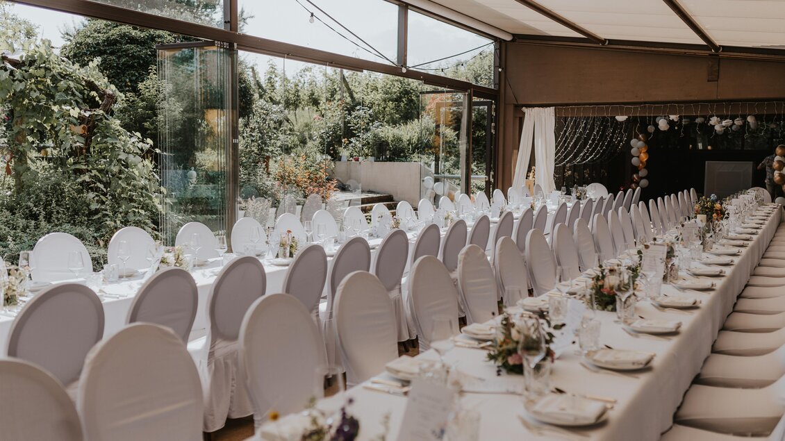 Ein eleganter Speisesaal mit langen weißen Tischen und Stühlen. Der Raum hat große Fenster mit Blick auf einen Garten. | © Weingartenhotel Harkamp