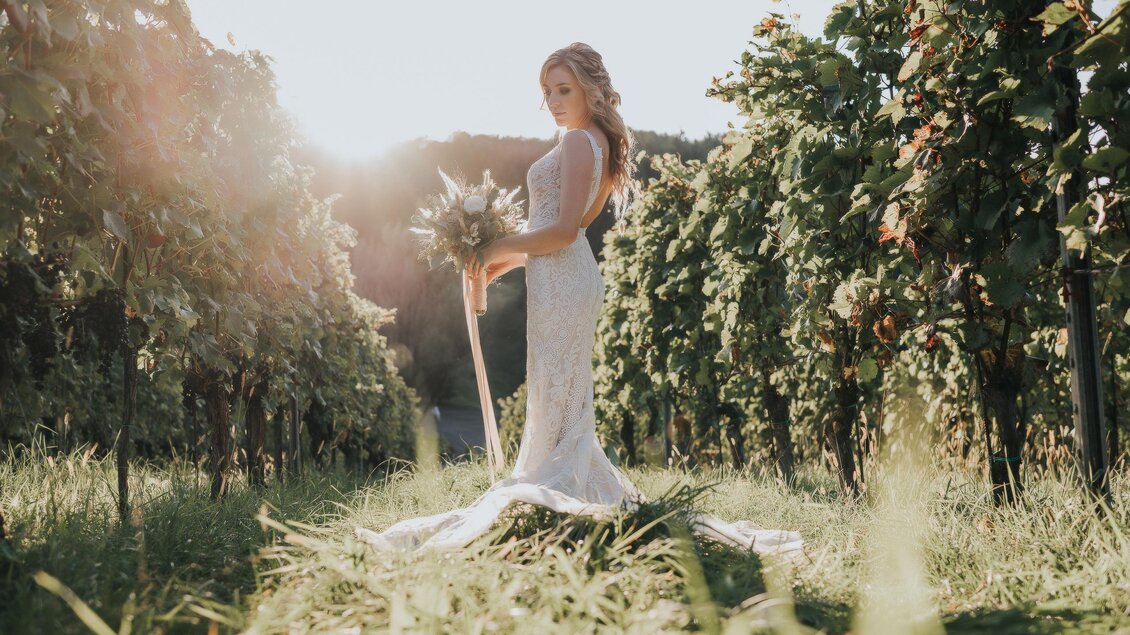 Eine Braut in einem traumhaften Hochzeitskleid steht in einem Weinberg. Die Sonne strahlt hinter ihr und schafft eine romantische Atmosphäre. | © Daniel Nagler | Weingartenhotel