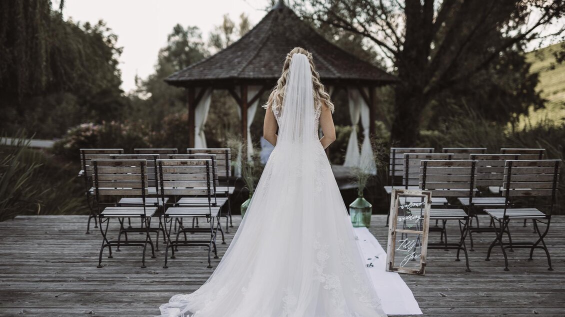 Eine Braut in einem langen weißen Kleid steht vor einem Pavillon. Reihen von Stühlen sind bereit für die Zeremonie im Freien. | © Weingut Georgiberg | Christine Wiener