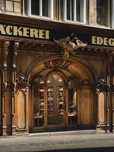 Historische Fassade der Hofbäckerei Edegger-Tax mit beleuchteten Schaufenstern und Eingang in Graz. | © Bernd Grosseck