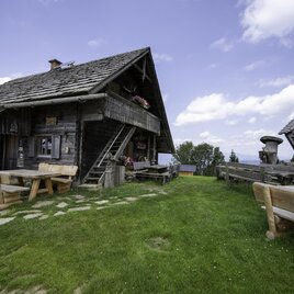 Eine gemütliche Berghütte mit einem gepflegten Garten und Holzterrassen. Im Hintergrund sind grüne Wiesen und eine teilweise bewölkte Himmelslandschaft zu sehen. | © Oststeiermark Tourismus