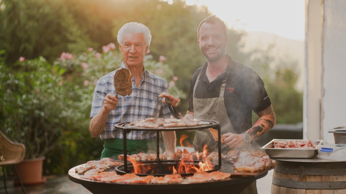 Zwei Männer grillen zusammen im Semriacherhof, einer hält ein großes Stück Fleisch, während der andere lächelt. Im Hintergrund brennt das Grillfeuer, und verschiedene Fleischstücke sind auf dem Grill. | © Wolfgang Hummer