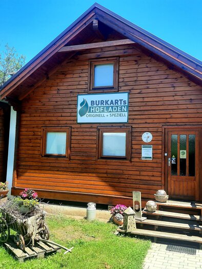 A rustic wooden house with a sign that says "Burkarts Hofladen". In front of the entrance, there are flower pots and an old wagon wheel decoration. | © Sigrid Burkart