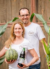 A couple stands between tall plants holding a melon and a bottle. The background features a wooden wall that conveys a rural atmosphere. | © Region Graz - Mias Photoart