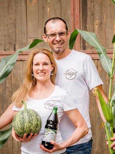 A couple stands between tall plants holding a melon and a bottle. The background features a wooden wall that conveys a rural atmosphere. | © Region Graz - Mias Photoart