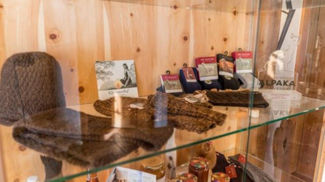 Eine Glasvitrine mit handgefertigten Wollartikeln wie Mützen, Schals und Socken. Im Hintergrund sind auch Gläser mit Honig und anderen lokalen Produkten zu sehen. | © Klausnerhof