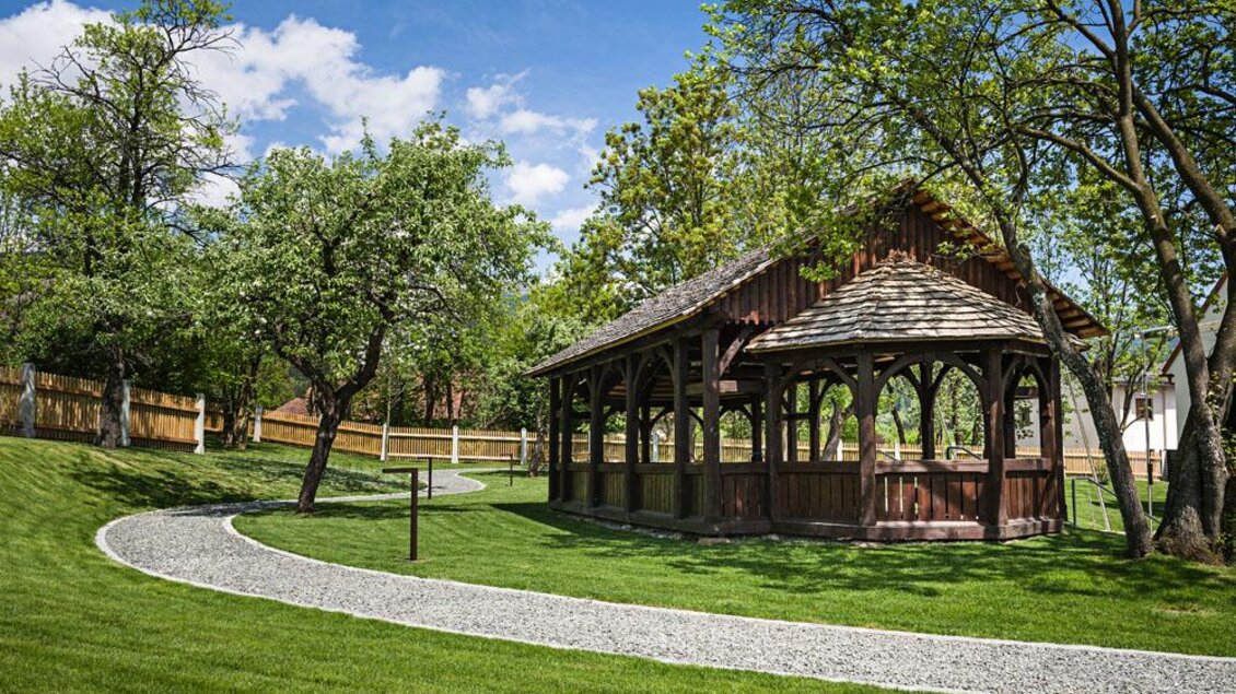 Ein schöner Garten mit einer überdachten Holzpergola und einer geschwungenen Kiesstraße. Umgeben von grünen Bäumen und herrlichem blauem Himmel. | © Tauroa GmbH