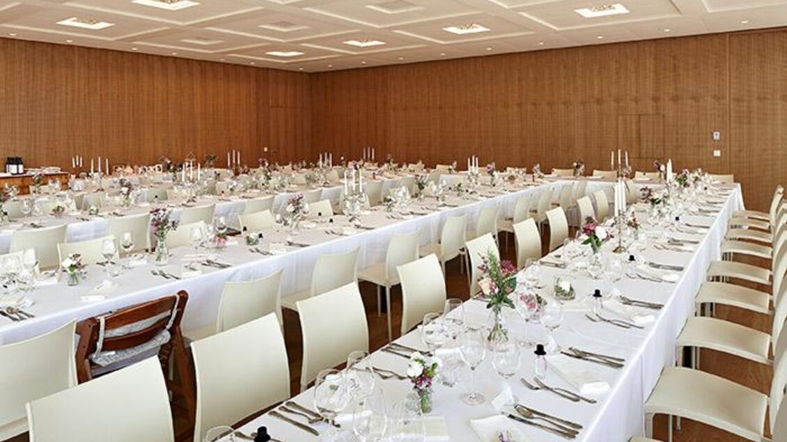 Ein eleganter Raum mit langen Tischen, die festlich gedeckt sind. Die Tische sind mit Blumen und Kerzen dekoriert, umgeben von hellen Stühlen. | © Tauroa GmbH