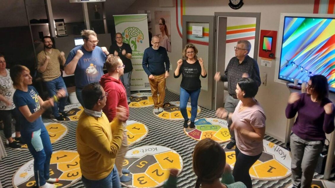 Eine Gruppe von Menschen tanzt und bewegt sich in einem hell erleuchteten Raum. Die Atmosphäre ist fröhlich und aktiv, mit bunten interaktiven Elementen im Hintergrund. | © Sabines Glücksplatz_Holzer