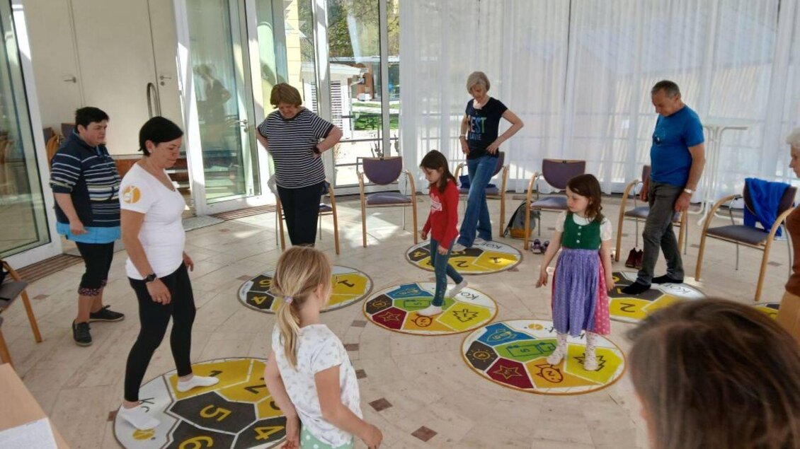Eine Gruppe von Kindern und Erwachsenen spielt ein Bewegungsspiel auf einem bunten Spielboden. Die Atmosphäre ist lebhaft und freundlich. | © Sabines Glücksplatz_Holzer