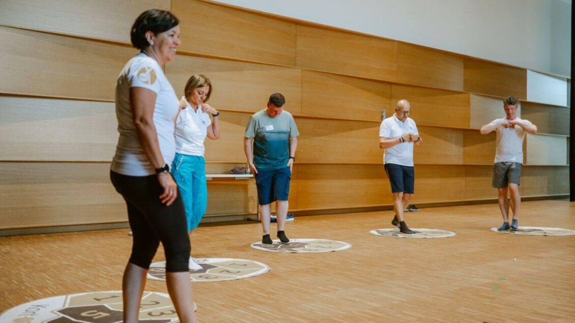 Eine Gruppe von fünf Personen steht in einem hellen Raum und interagiert auf großen runden Matten. Alle tragen sportliche Kleidung und scheinen an einer Aktivität teilzunehmen. | © Sabines Glücksplatz_Holzer