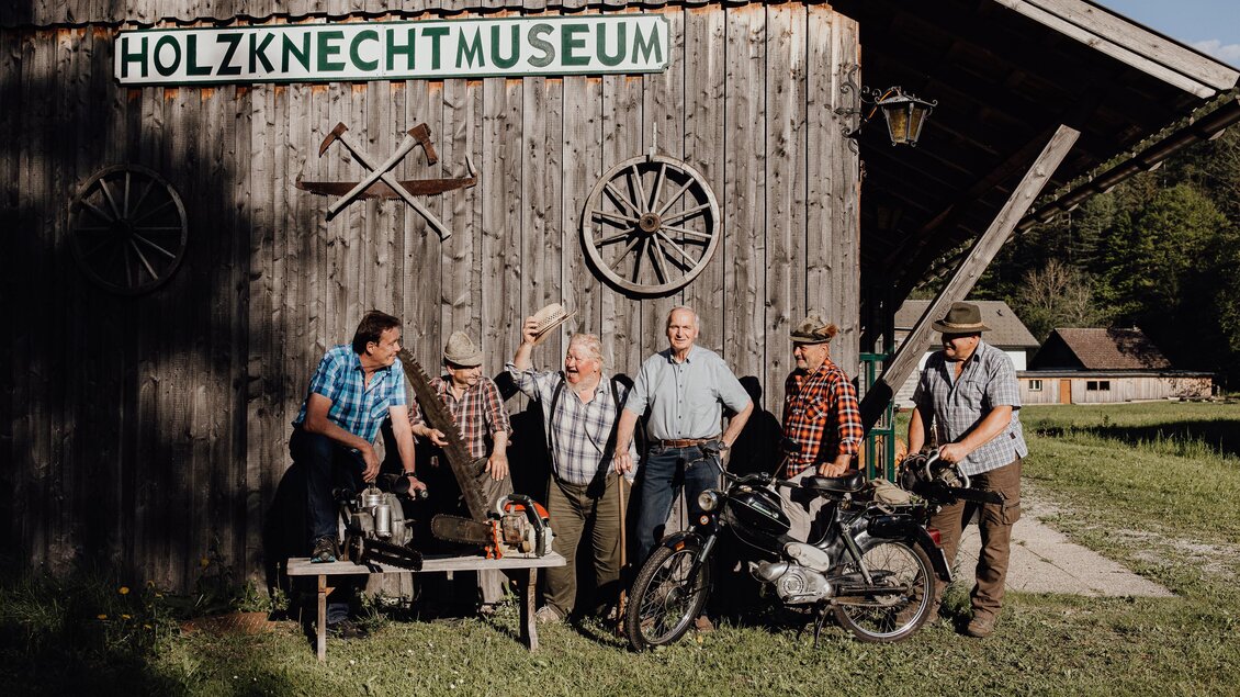 Eine Gruppe von Männern steht vor dem Holzknechtmuseum. Sie tragen traditionelle Kleidung und posieren mit Werkzeugen und einem Motorrad. | © nicoleseiser.at