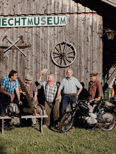 Eine Gruppe von Männern steht vor dem Holzknechtmuseum. Sie tragen traditionelle Kleidung und posieren mit Werkzeugen und einem Motorrad. | © nicoleseiser.at