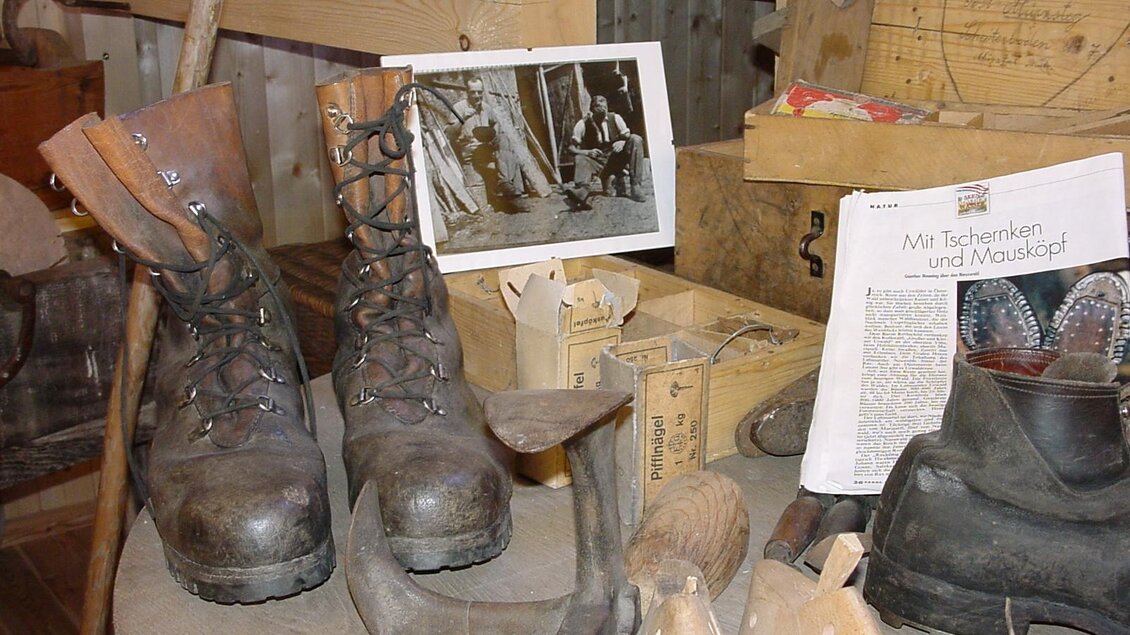 Ein historischer Arbeitsplatz mit alten Stiefeln, Werkzeugen und fotografischen Erinnerungen. Auf dem Tisch sind auch einige Zeitungsartikel und handwerkliche Materialien zu sehen. | © Holzknechtmuseum