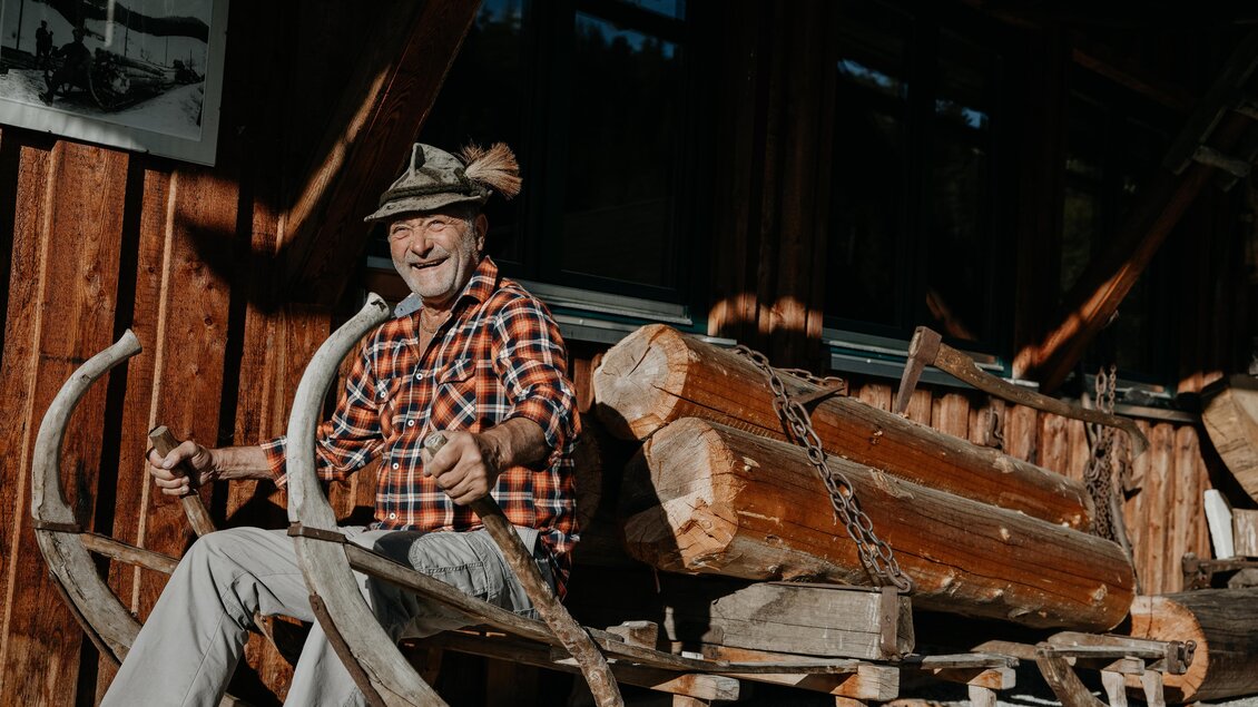 Ein älterer Mann sitzt auf einem Holzschlitten und lächelt freundlich. Im Hintergrund sind Holzarbeiten und eine Holzfassade zu sehen. | © nicoleseiser.at