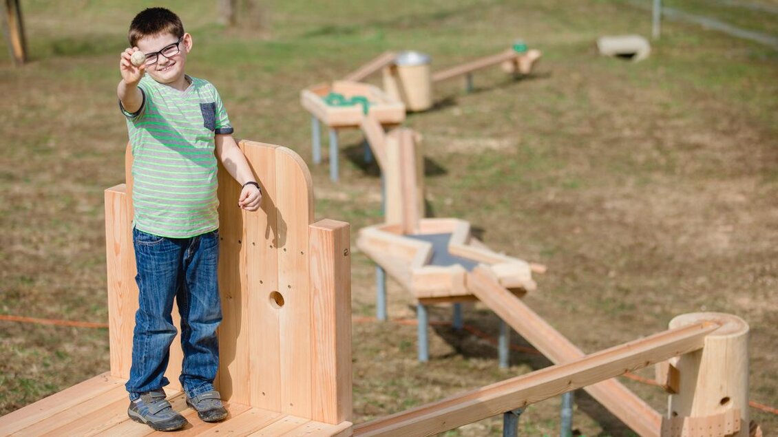 Ein Junge steht auf einem Holzspielplatz und zeigt mit dem Finger. Im Hintergrund sieht man verschiedene Holzstrukturen zum Spielen. | © Karin Bergmann