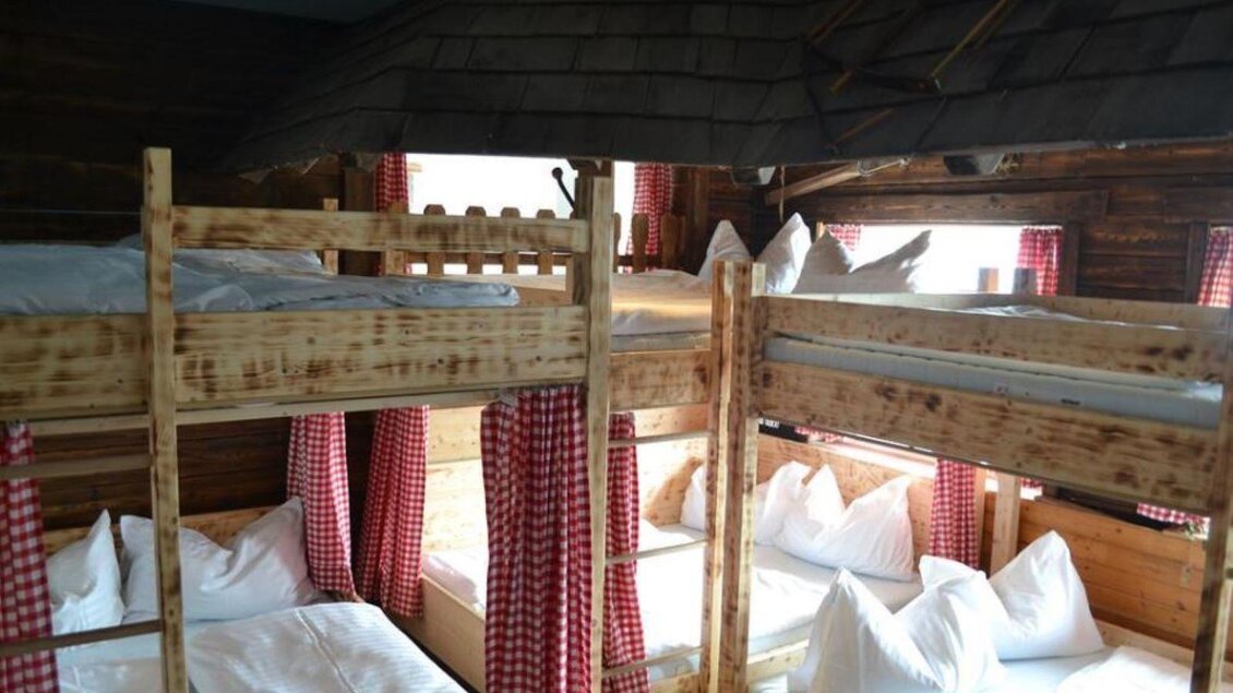 Ein gemütlicher Schlafraum mit Etagenbetten aus Holz. Die Fenster sind mit rot karierten Vorhängen geschmückt und es gibt mehrere Kopfkissen auf den Betten. | © Hoo-Ruck Alm