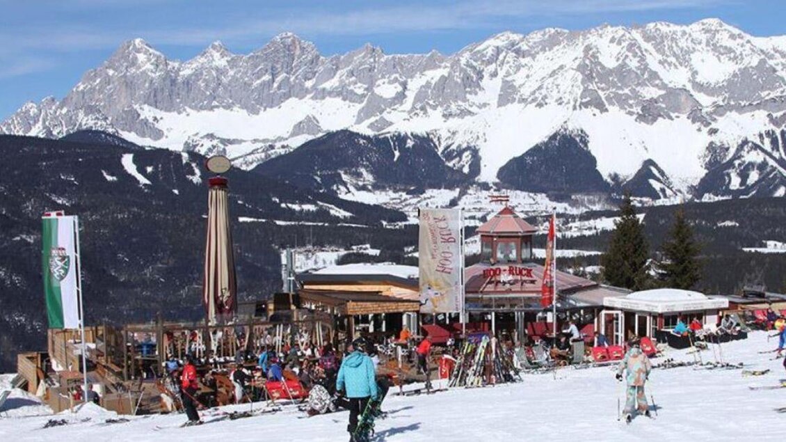 Ein lebhaftes Skigebiet mit einem rustikalen Restaurant im Vordergrund. Im Hintergrund erheben sich majestätische, schneebedeckte Berge unter einem klaren Himmel. | © Hoo-Ruck Alm
