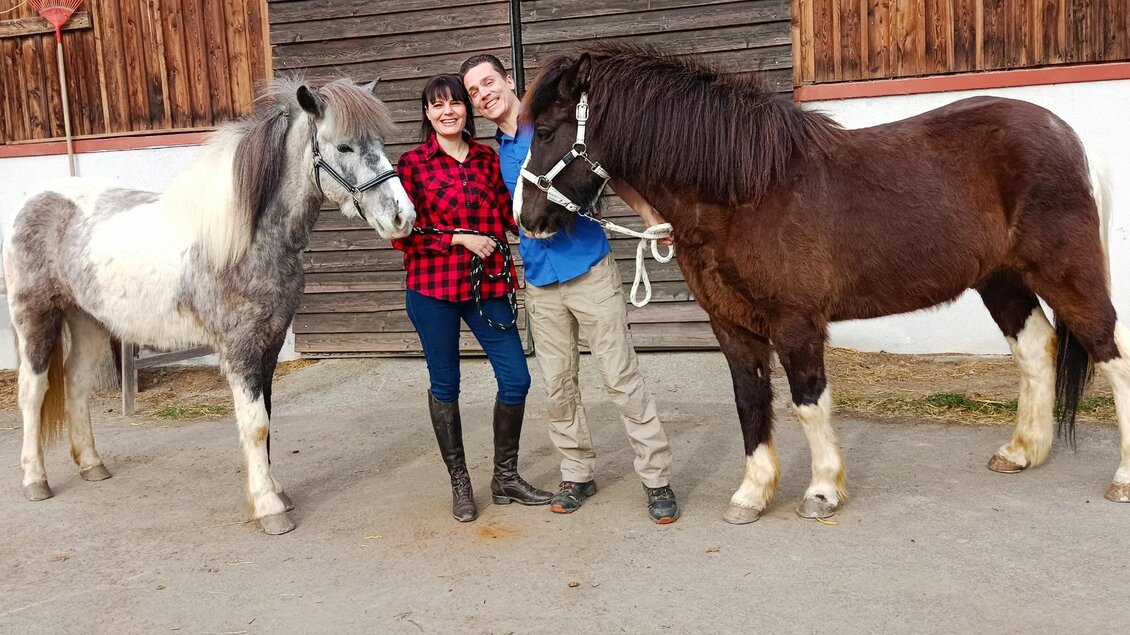Eine Frau und ein Mann stehen lächelnd zwischen zwei Pferden. Sie befinden sich vor einer Holzscheune in einem ländlichen Umfeld. | © Hopp Galopp