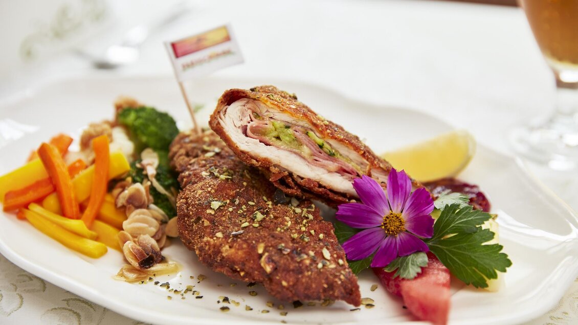 Ein köstliches Gericht auf einem Teller, das paniertes Fleisch mit Schinken und Gemüse präsentiert. Als Garnitur sind frische Kräuter und eine Blume dekorativ angeordnet. | © Klöcherhof Domittner