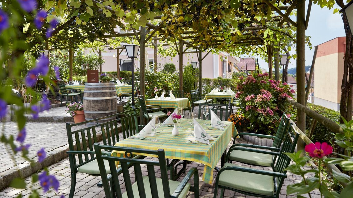 Ein einladendes Restaurant im Freien mit grünen Stühlen und Tischen unter einem schattigen Dach aus Pflanzen. Bunte Blumen und eine entspannte Atmosphäre schaffen einen schönen Essbereich. | © Klöcherhof Domittner