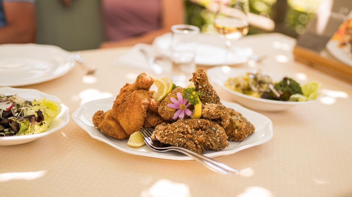 Ein Teller mit knusprigem Hähnchen, dekoriert mit einer Blume und Limetten. Im Hintergrund sind mehrere Salate und Gläser zu sehen.