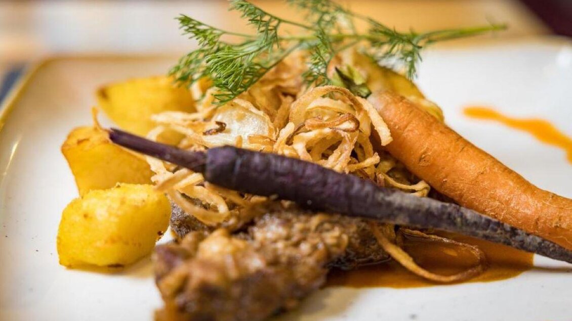 Ein kunstvoll angerichtetes Gericht mit gebratenem Fisch, knusprigen Zwiebelringen und buntem Gemüse. Beilagen sind goldene Kartoffeln und eine Karotte. | © Lindenhof