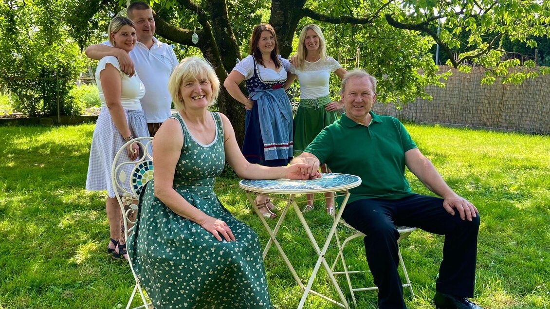Eine Gruppe von sechs Personen posiert im Freien auf einer Wiese unter einem Baum. Zwei Personen sitzen an einem kleinen Tisch, während die anderen im Hintergrund stehen. | © Hotel & Wirtshaus Hödl-Kaplan