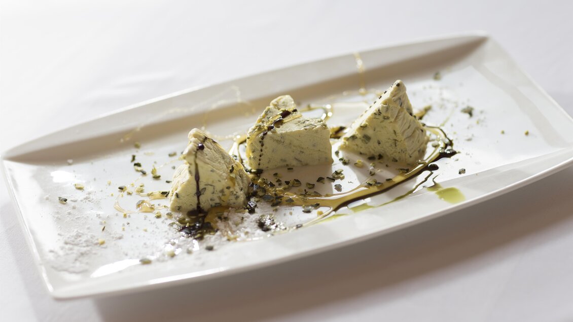 Ein elegantes Dessert auf einem weißen Teller, bestehend aus dreieckigen Stückchen. Die Garnitur umfasst Kräuter und eine dunkle Sauce. | © Karl Schrotter
