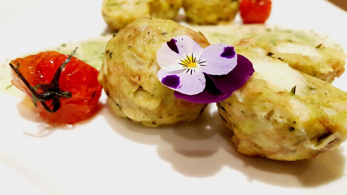Ein Teller mit köstlichen, kleinen Knödeln, garniert mit einer essbaren Blume. Neben den Knödeln liegen gegrillte, rote Tomaten. | © Teuschler Mogg