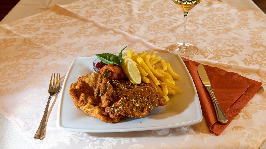 Ein Teller mit paniertem Fleisch, Pommes Frites und Zitronenscheiben. Daneben steht ein Glas Wein auf einem Tisch mit einer eleganten Tischdecke. | © Hotel Neuhold