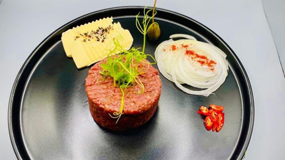 Ein elegantes Gericht mit rohem Fleisch, garniert mit frischen Kräutern. Neben dem Fleisch liegen Käse, Zwiebeln und eine kleine Chili. | © Hotel-Restaurant Schwarzer Adler