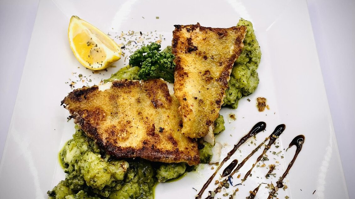 Ein Teller mit zwei knusprigen Fischfilets auf einer cremigen grünen Beilage. Mit einem Stück Zitrone und kunstvollen Sauce-Dekoren serviert. | © Hotel-Restaurant Schwarzer Adler