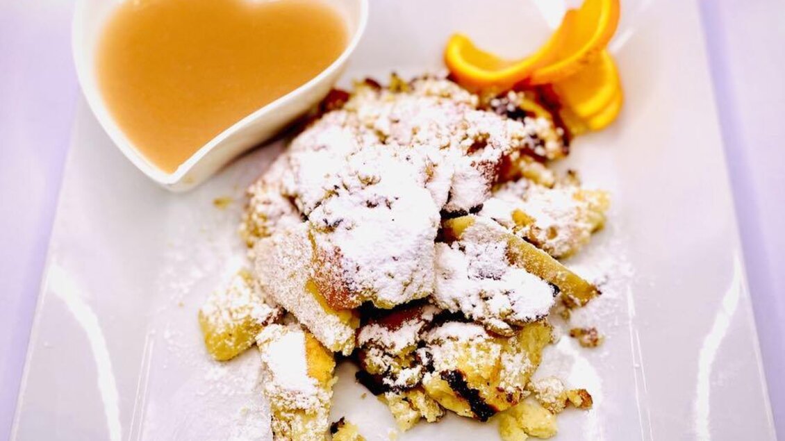 Ein Teller mit köstlichem Dessert, das aus kleinen Stückchen gebackenem Brot mit Puderzucker und frischen Orangenstücken besteht. Dazu gibt es eine herzförmige Schüssel mit Sauce. | © Hotel-Restaurant Schwarzer Adler